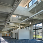 An interior shot of the newly renovated train building with blue flooring an exposed white ceiling and an office with white walls in the center of the photo.