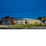 The eastern view of the newly renovated trainyard building with interior lights on on a cloudy evening.