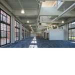 Interior view of the newly renovated train building with blue flooring and exposed white ceiling and natural light shining through the large windows. 