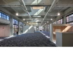 Interior view overviewing the front entrance of the newly renovated train building with blue flooring and exposed white ceiling and natural light shining through the large windows. 
