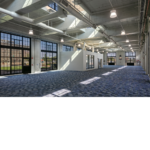Interior side view of the newly renovated train building with blue flooring and exposed white ceiling and natural light shining through the large windows. And an office with white exterior walls inside the building.