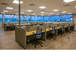 Two rows of cubicles with grey walls and light wooden desks with gray and blue chairs tucked in.