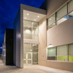 A well-lit-up view of the side entrance of the Chevron Field Office with a window wall showing the well-lit-up interior of the inside of the building. 