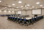 angle shot of a large room with two separate groups of tables and chairs pointing toward a large white screen and a projector hanging from the ceiling. 