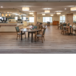 Cafeteria with neatly set up tables and wooden chairs with green cushions.