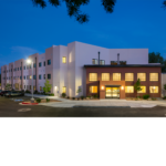 Nighttime photo of the La Vida Llena building. Windows are visible on the white wall and glowing interior lights shine from the entrance building.  