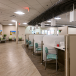 Check-in cubicles with teal chairs and a waiting area to the left with green chairs and pictures hanging on the wall. 
