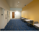 Hallway with blue carpet and yellow wall with desks on lined up against the wall.